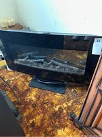 Front view of the black glass electric fireplace with faux logs inside sitting on a floor with a patterned carpet.