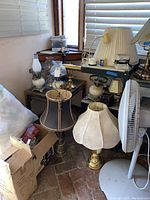 Photo showing multiple lamps with various shades grouped around the end table. Includes some boxed items and a fan.