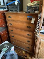 Front view of tall boy dresser showing five drawers with ornate dark metal handles, placed in a room with other furniture.