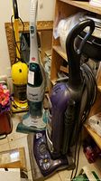Photo showing two Eureka vacuum cleaners and one Hoover FloorMate cleaner along with mops and brooms in a cluttered corner