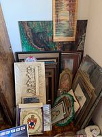 Photo showing a collection of various framed items leaned against wall, includes wall plaques, framed canvas art pieces, and mirrors of various sizes and styles.
