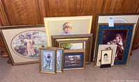 Full view of 8 assorted framed vintage wall art pieces grouped on floor: large oval orchard scene, floral prints, portraits of children, and small decorative scenes.