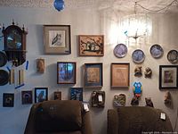 Wide view of wall display with multiple framed owl pictures, collector plates, carved owl plaques and vintage portraits on a beige wall above two brown chairs.