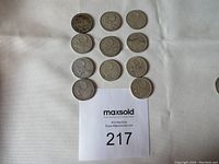 Top-down view of 11 Canadian silver quarters arranged on a white cloth, showing both obverse and reverse sides with a MaxSold lot number card present.