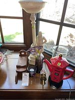 Photo showing entire lamp, red teapot kettle, vases with flowers, candle holder, and wooden handled item on wood surface in front of window lighted by daylight.