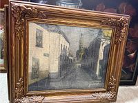 Framing and artwork showing the street scene painting with aged canvas crackling and brown ornate carved floral corners of the frame.