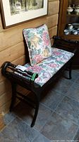 Photo of a wooden bench with dark finish showing the floral upholstered seat cushion and matching throw pillow against a wood-paneled wall and tile floor.