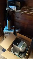 Photo showing a Breville coffee maker in a box, Hamilton Beach percolator box, stainless steel coffee dispenser, and other items on a shelf.