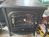 Black metal faux stove air heater, model 6901050100, showing front glass panel with simulated logs and brass handle.