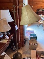 Full view of vintage floor lamp with beige fabric shade and bronze/brass metal pole next to a wooden wall and other lamps.
