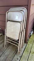 Stack of 4 beige metal folding chairs leaning against exterior siding on a wooden deck. Chairs show surface rust and paint wear.