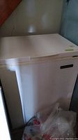 Front and left side view of white chest freezer with dents and scrapes, with a plastic bag on floor nearby.