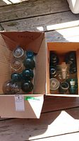 Two cardboard boxes containing assorted glass insulators in green, teal, blue, and clear colors with some showing chip damage and cracks.