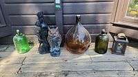 Wide view of four glass jugs in green, amber, olive colors, a black plastic owl statue, a black decorative plastic statue, and small black metal bird feeder lantern on weathered wooden surface.