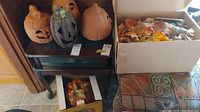 Three jack-o'-lantern style ceramic pumpkins (two orange, one gray) and one large plain orange ceramic pumpkin on shelves. Boxes with artificial fall leaves and witch decorations to the side.
