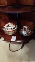Covered dish with lid and decorative stand next to tea cups and saucers on wooden shelf.