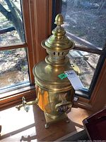 Full view of the brass-toned samovar positioned on a wooden surface near a window, showing entire height and design.