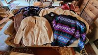 Photo showing six folded hand knit wool sweaters and cardigans including cream cable knit and colorful pattern designs.