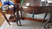 Two wooden tables shown, a larger curved front side table and a smaller straight-legged side table, both made of quarter sawn tiger oak with visible tiger stripe grain pattern. The larger table has brass ring drawer pulls and a green tassel on drawer keyhole.