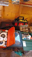 Wide shot of Husky tool carrier, Skil jigsaw bag, orange metal box with polishing wheels, green toolbox with drill bits, and other small tools on wooden bench