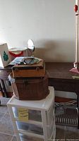 View of white plastic storage cart with three drawers on casters; brown woven-pattern plastic sewing box; wooden carrying case and partial view of Singer cabinet behind.