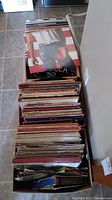 Long overhead view of cardboard box filled with ~60 vinyl record sleeves, two rows. Floor tile background.