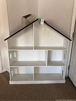 Front view of the white doll house shaped bookcase showing 6 compartments with black roof edges and some visible marks on the wood.