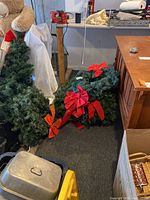 Photo of several artificial Christmas wreaths leaning against wall and floor, including one larger and multiple smaller wreaths, all with red bows attached