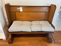 Front view of wooden church pew bench with beige cushion on seat, showing armrests and high backrest.