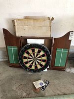 Photo showing the dartboard, its wooden wall case with open side doors, and darts with accessories on the floor in front.