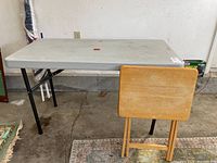 Lifetime white plastic folding table with black legs alongside a wooden TV tray, both showing signs of use and dust.