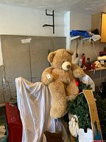Large light brown plush giant teddy bear seated on a covered surface with part of the bear visible and surrounding items in a garage environment.