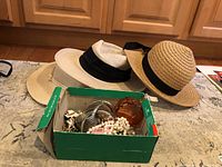 Three hats in front of kitchen cabinets, two straw hats and one white mesh hat with black bands, with a green box of costume jewelry containing various bracelets, necklaces, and pins.