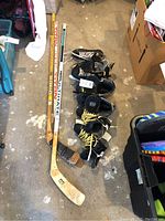 Photo showing four pairs of hockey skates aligned on floor with three hockey sticks beside them.