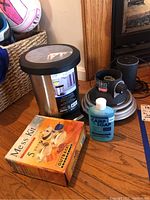 Photo showing all items in the lot on wooden floor: 5 piece mess kit box, stainless steel step can, propane heater, Bluetooth speaker, and Camp Soap bottle.