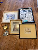Photo showing six picture frames on the floor, including a framed historic newspaper clipping, maritime scene, small gold-tone frame with black and white print, and various other framed artworks.
