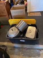 Plastic bin containing multiple metal roasting pans, Dutch ovens, pots, and lids, some stacked inside others, showing wear and used condition. Yellow bin lid open above cookware.
