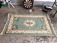 Photo of first Almora oriental style rug laid flat on concrete floor. Green background with cream and gold border, floral accents, frayed fringed edges.