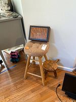 Larger stool with woven seat, smaller milking stool, and wooden box displayed together on wood floor against white wall.