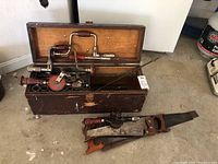 Closed view of wooden toolbox filled with antique hand tools including drills, screwdriver, wrench, and three hand saws beside it