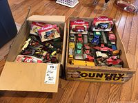 Two cardboard boxes filled with various vintage toy trucks, including three Campbell's 100th Anniversary die cast trucks in original packaging, and assorted loose vintage toy vehicles.