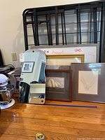 Lots of wall art pieces and mirror arranged on floor including calendar and framed prints; larger frame with ship drawing has broken glass