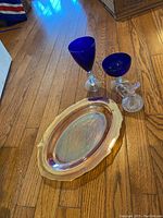 Two cobalt blue wine glasses, one cobalt blue champagne glass, small clear glass pitcher, and iridescent Federal glass oval plate placed on wood floor.