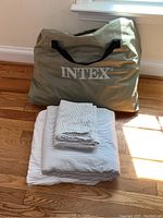 Beige INTEX carry bag containing the folded air mattress with folded striped linens in front.