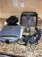 Photo of the full set of appliances including the hand mixer with attachments, sandwich maker, waffle maker, and toaster lined up on a granite counter.