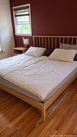 Full view of king size wooden platform bed frame with slatted headboard, white linens, mattress visible, hardwood floor and window in background.