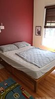 Full view of the full size bed frame with mattress, pillows, fitted sheet and comforter against a red wall with window.