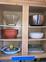 Shelf with various mixing bowls: large clear glass, stainless steel, textured orange plastic, and white ceramic. Also glass bakeware dishes and citrus juicer parts on lower shelf.