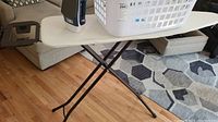 White ironing board with black metal legs with Black & Decker iron and plastic laundry basket on top, shown on wooden floor in living room