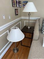 Two table lamps placed on a wood floor next to a dark side table and a beige couch. Both lamps have white fabric shades and brushed metal bases.